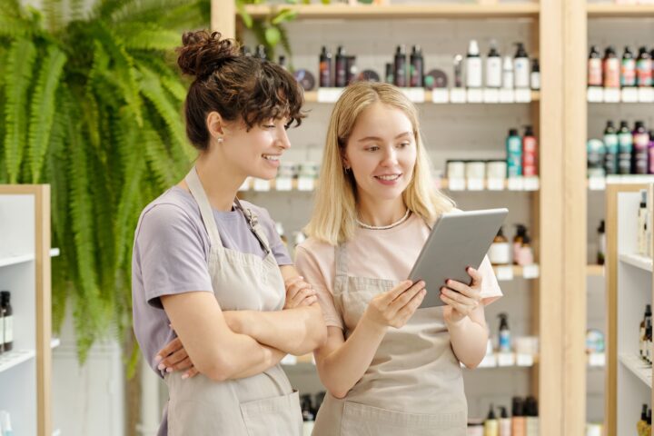 two-young-smiling-shop-assistants-looking-at-table of pos system in the cosmetic and beauty store